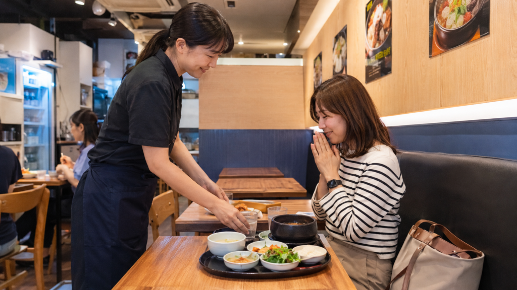 韓国で一人でも入りやすい店に入り、定食を安心して楽しむ日本人女性旅行者と親切なスタッフの様子